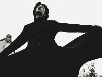 Model in a long black coat captured from a low angle with arms outstretched against a bright sky.