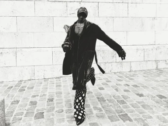 Black man dancing in a long dark coat and polka dot trousers outdoors against a stone block wall for a HERO Magazine editorial project.