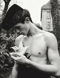 Monochrome portrait of a topless man wearing a dark patterned cap, holding a white lily flower outdoors.
