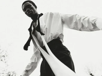 Low-angle monochrome fashion editorial portrait of a young black man wearing a white shirt and dark trousers against a plain sky.