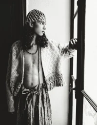 Model with long dark hair wearing a patterned jacket and beaded cap standing by a window for HERO Magazine.