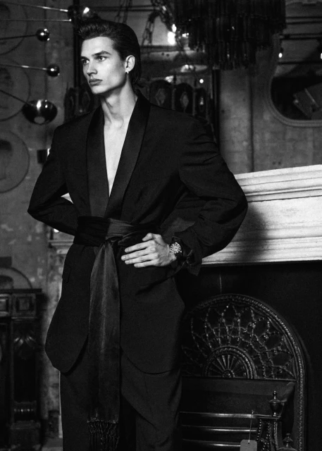 Black and white portrait of a male fashion model posing indoors wearing a belted black suit. High contrast and shallow depth of field.