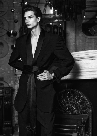 Black and white portrait of a male fashion model posing indoors wearing a belted black suit. High contrast and shallow depth of field.