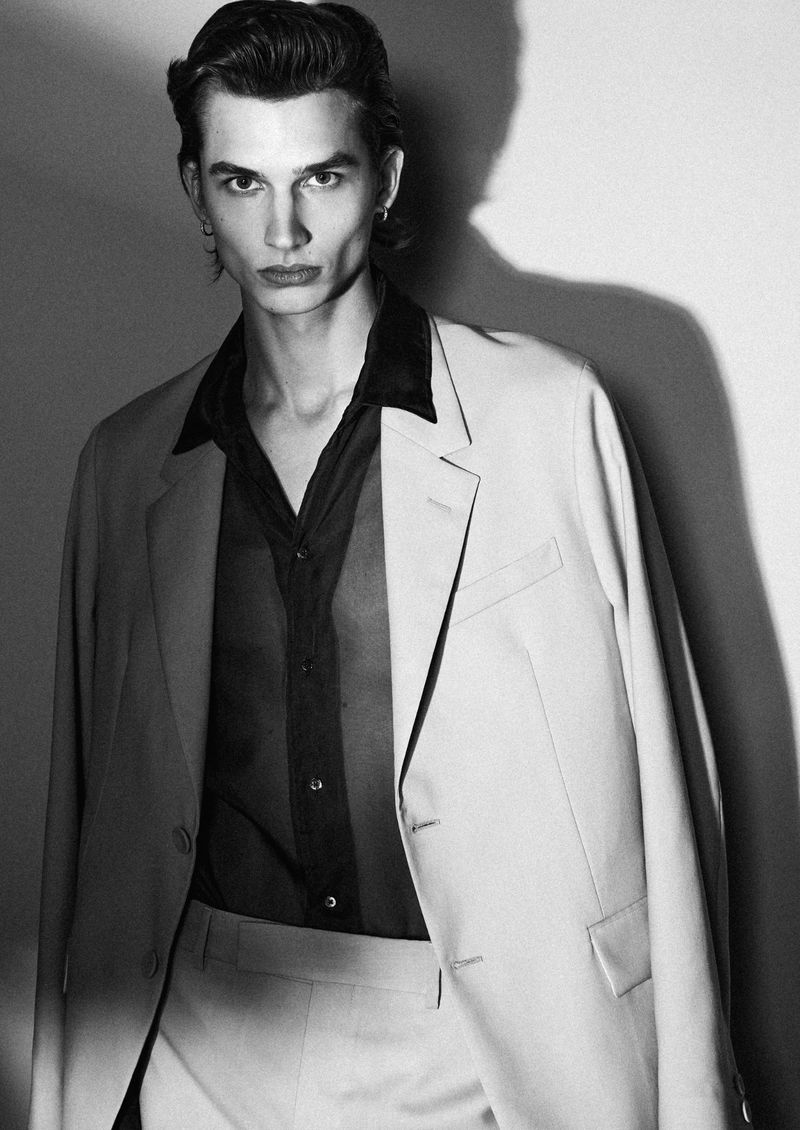 Monochrome portrait of a male model in a suit jacket and dark shirt against a plain studio wall with harsh shadows.