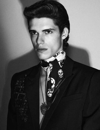 Monochrome portrait of an adult male model wearing a black suit, skull-print scarf, and pendant necklace in a studio setting.