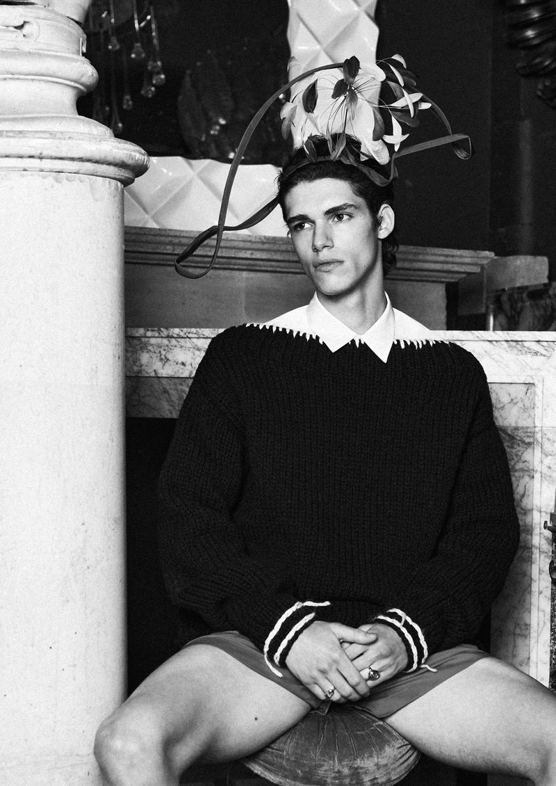 Black and white portrait of a male model in a dark sweater with white accents, sitting indoors with decorative objects in the background.