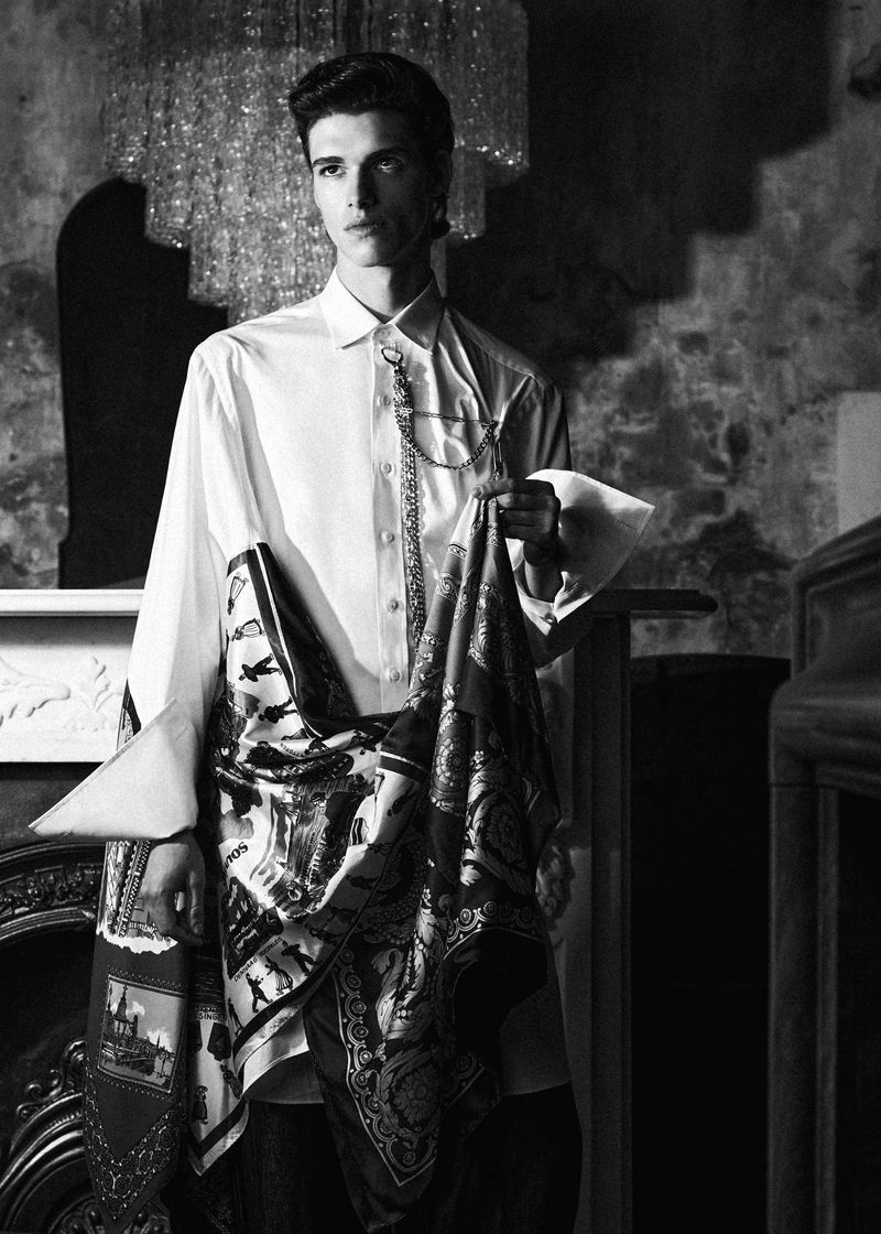 Monochrome fashion portrait of a male model wearing a white button-down shirt and draped patterned scarf standing indoors