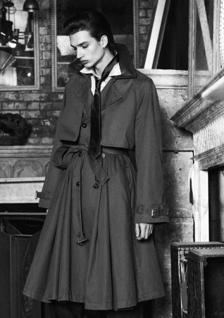 Monochrome fashion editorial portrait of a young man wearing a belted trench coat and tie standing indoors by a fireplace