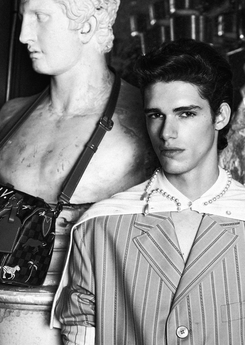 Monochrome portrait of a young adult male model wearing a striped blazer and pearl necklace posing next to a classical marble statue.