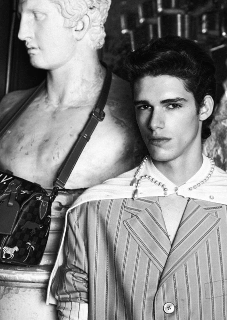 Monochrome portrait of a young adult male model wearing a striped blazer and pearl necklace posing next to a classical marble statue.