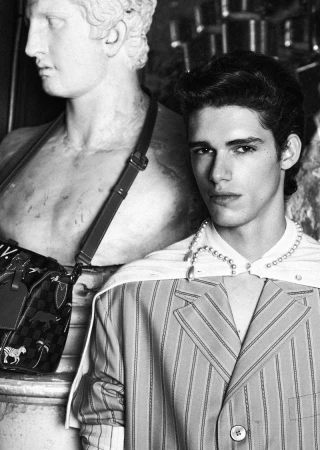 Monochrome portrait of a young adult male model wearing a striped blazer and pearl necklace posing next to a classical marble statue.