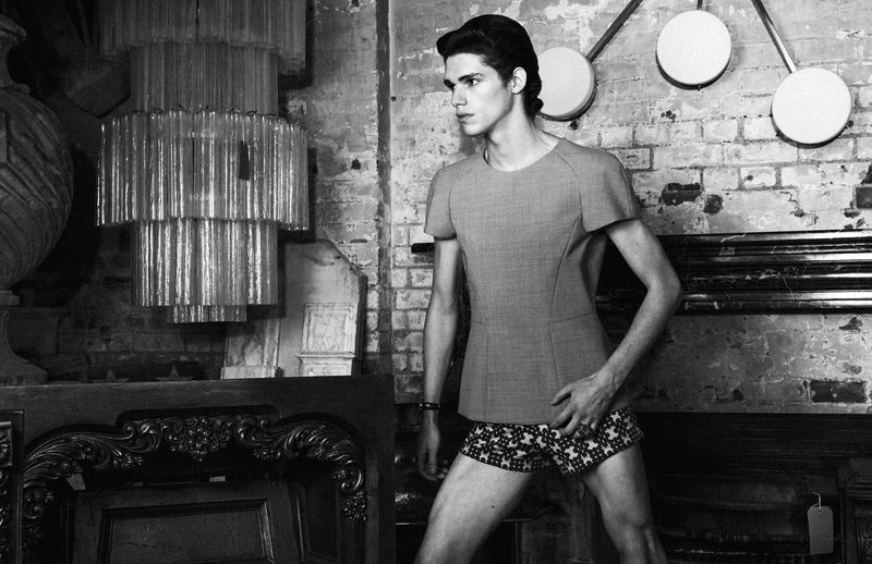 Black and white studio fashion shot of a slender adult male in a contemporary outfit, posing against a distressed brick wall backdrop.
