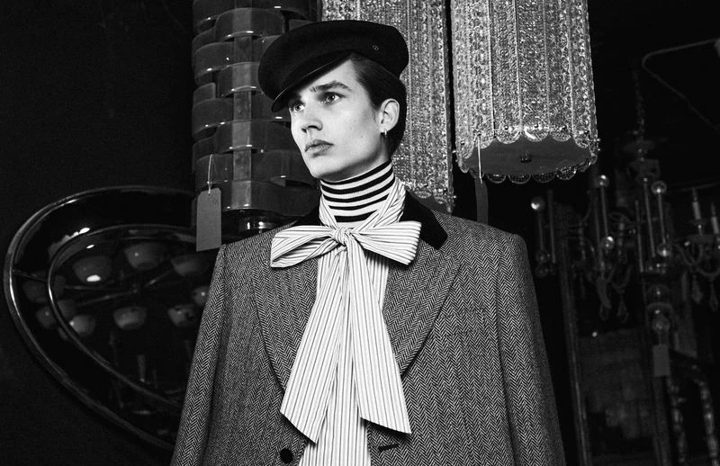 Monochrome portrait of a slender adult male in a retro herringbone suit, cap, and striped necktie, against an ornate background.