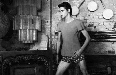 A young male model posing in an interior space with a large chandelier and exposed brick wall