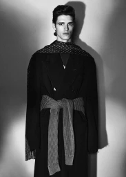 Monochrome portrait of a male model wearing a dark coat and tied scarf against a seamless background with dramatic shadow casting.