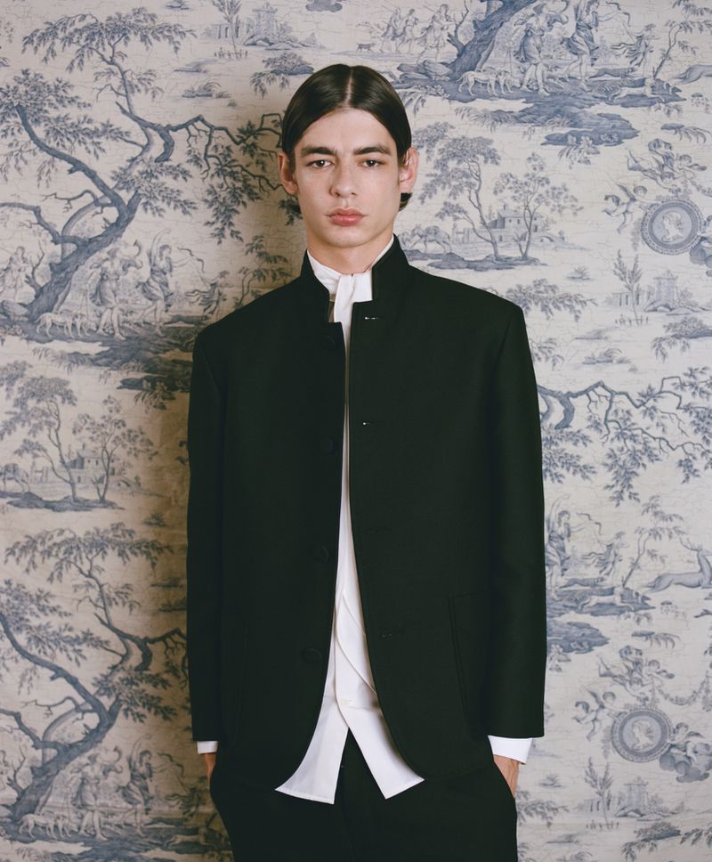 Portrait of a young adult man in a black jacket and white collared shirt against a blue-and-white toile-patterned wall.