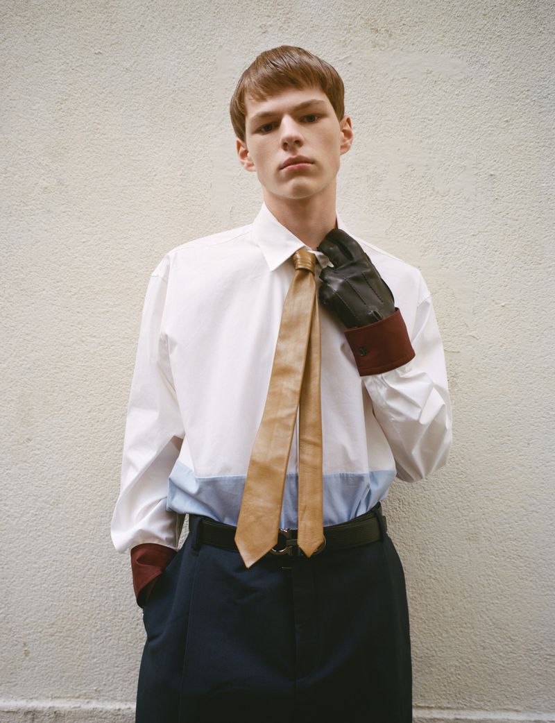 Young adult man in white shirt, tie, and gloves poses in front of a neutral wall, conveying a modern fashion aesthetic.