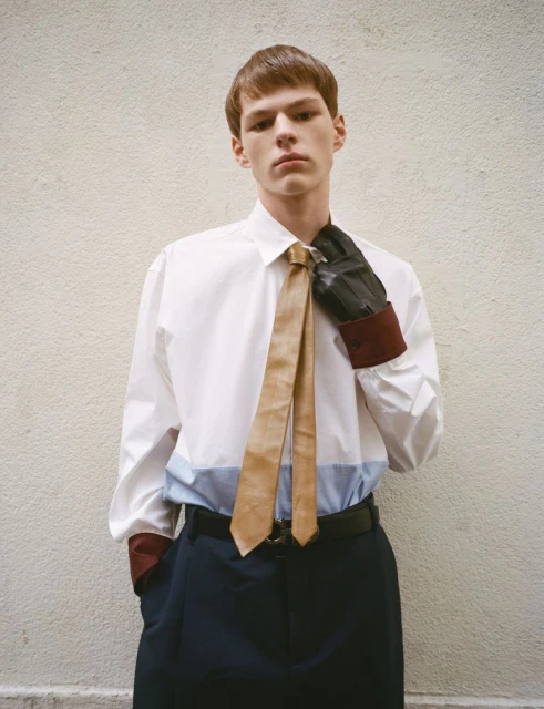 Young adult man in white shirt, tie, and gloves poses in front of a neutral wall, conveying a modern fashion aesthetic.