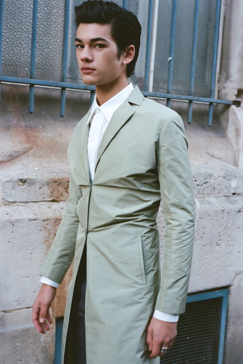 Young adult male in a light green trench coat stands against a textured wall and blue metal window frame, fashion lifestyle editorial.