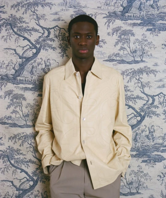 Portrait of a dark-skinned male model in light colored jacket and pants posing in front of a blue and white toile wallpaper indoors.