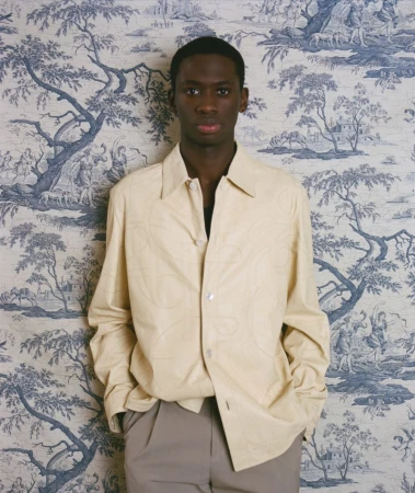 Portrait of a dark-skinned male model in light colored jacket and pants posing in front of a blue and white toile wallpaper indoors.