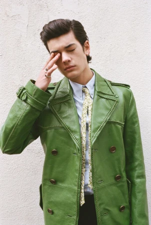 A young man with dark hair wears a bright green double-breasted leather coat over a pale blue shirt and patterned tie against a white wall.