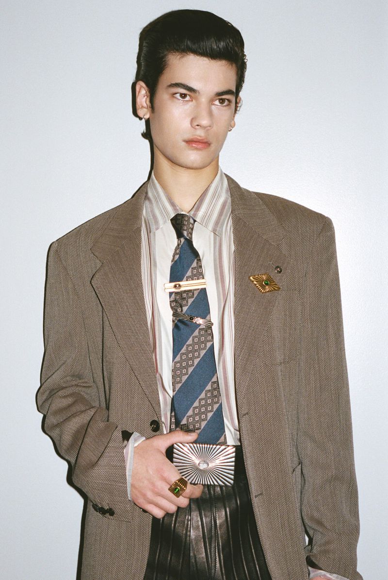 Portrait of a stylish male fashion model wearing a blazer, collared shirt, and tie while holding a metallic card holder in a studio.