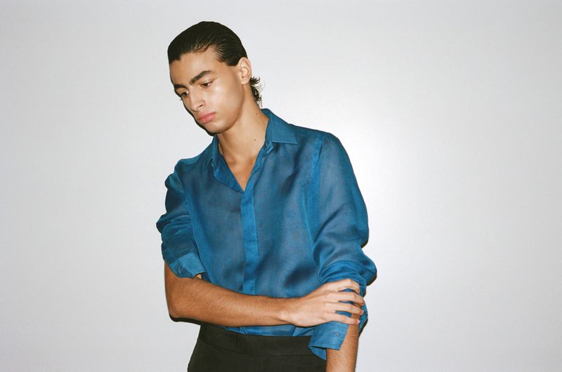 A man with slicked-back dark hair wearing a translucent blue button-up shirt and black trousers, posing against a white studio wall.