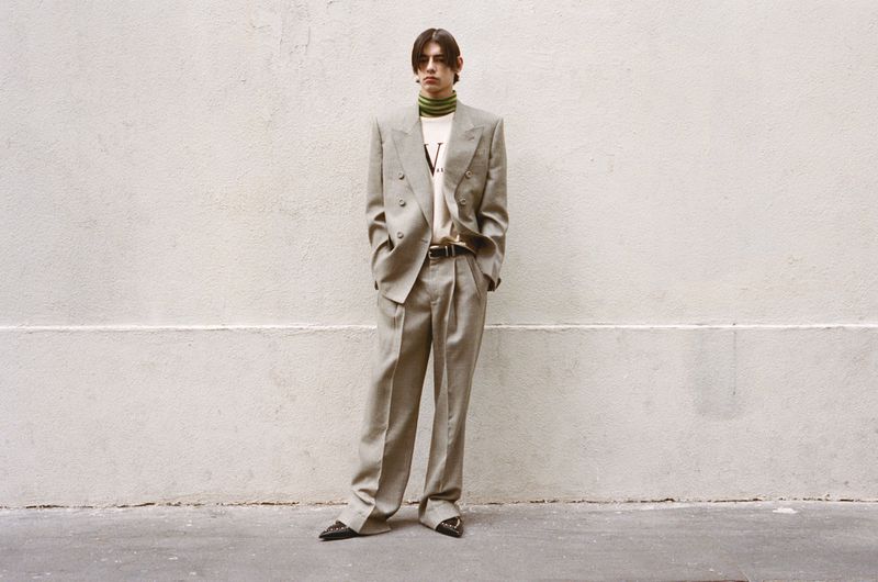 Full-length shot of a young adult male fashion model in a grey double-breasted suit, turtle-neck sweater, and black dress shoes against a wall.