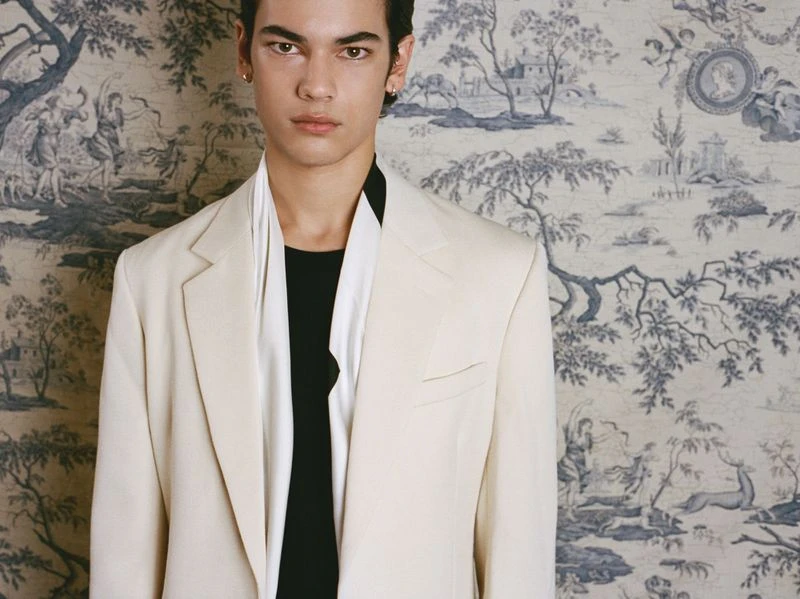 Portrait of an adult male wearing a cream-colored blazer and black shirt against a wallpaper backdrop featuring a toile pattern.