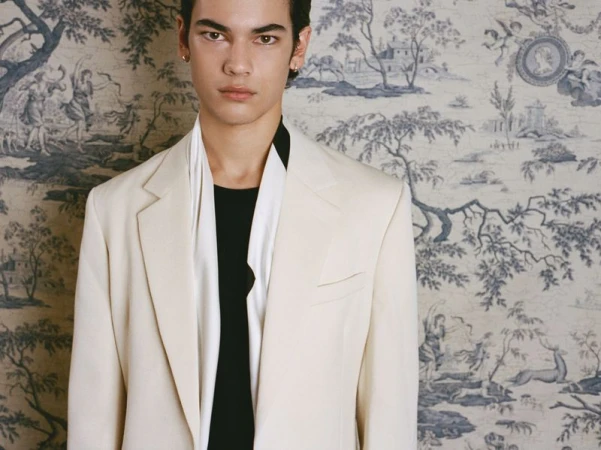 Portrait of an adult male wearing a cream-colored blazer and black shirt against a wallpaper backdrop featuring a toile pattern.