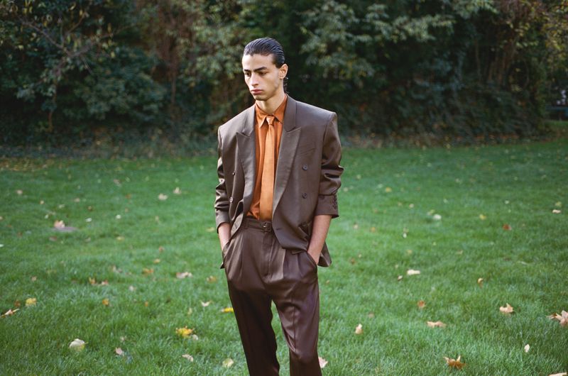 A young adult male in a brown suit stands on a green lawn in front of trees. He has his hands in his pockets in a full shot.