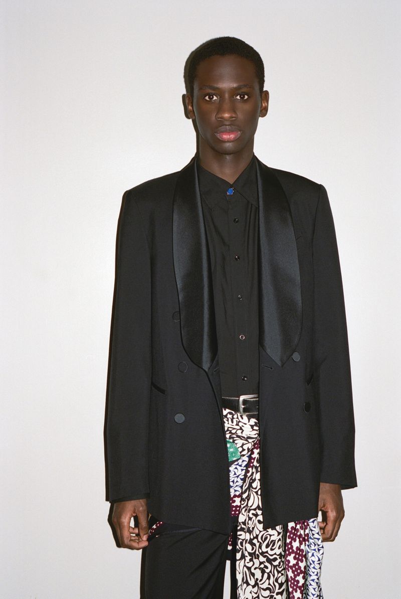 Man in black tuxedo and patterned skirt stands against white backdrop. High-fashion, striking pose, and unique blend of formal and informal.