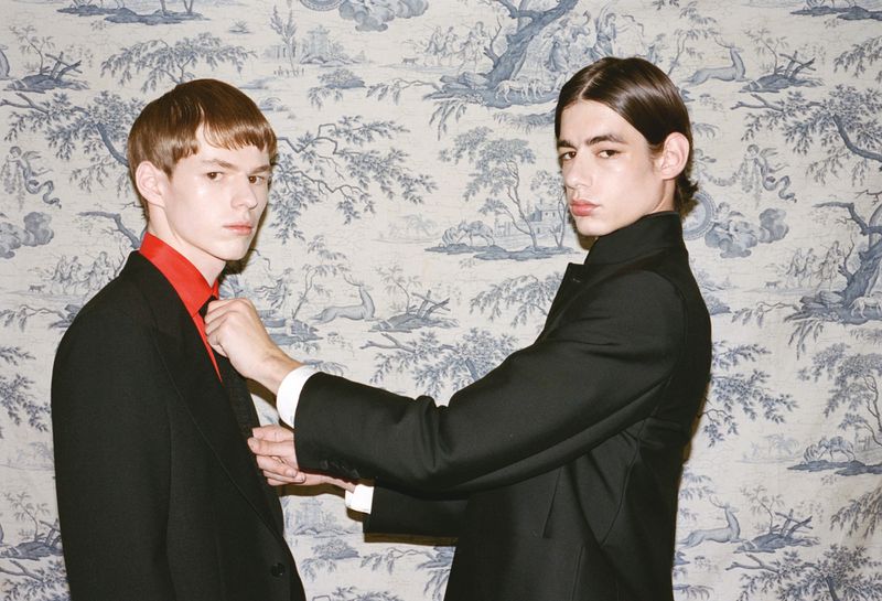 Two stylish young men in dark suits pose indoors against a patterned wallpaper. One man adjusts the other's red collared shirt.