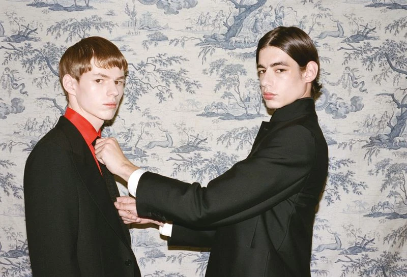 Two stylish young men in dark suits pose indoors against a patterned wallpaper. One man adjusts the other's red collared shirt.