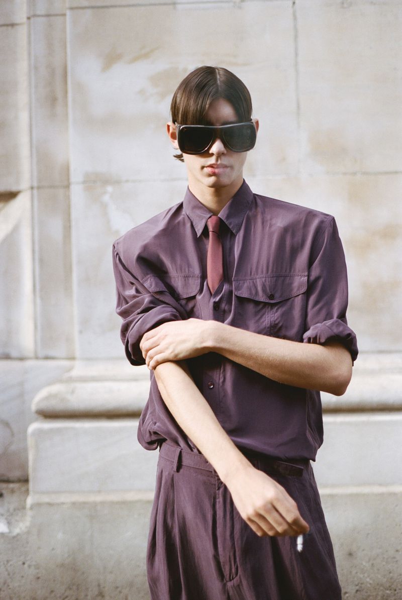 A stylish person wearing a purple shirt, tie, and sunglasses stands outdoors in a fashion-forward pose, exuding a cool, editorial aesthetic.