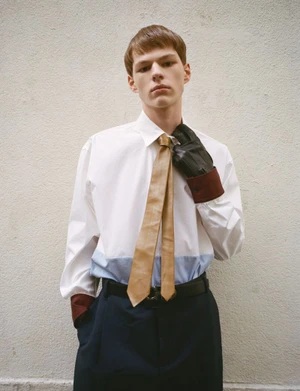 Male model in white shirt with tan tie and dark trousers standing against a textured wall for HERO Magazine