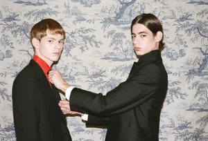 Two male models in dark suits standing against a patterned blue and white wallpaper background for HERO Magazine