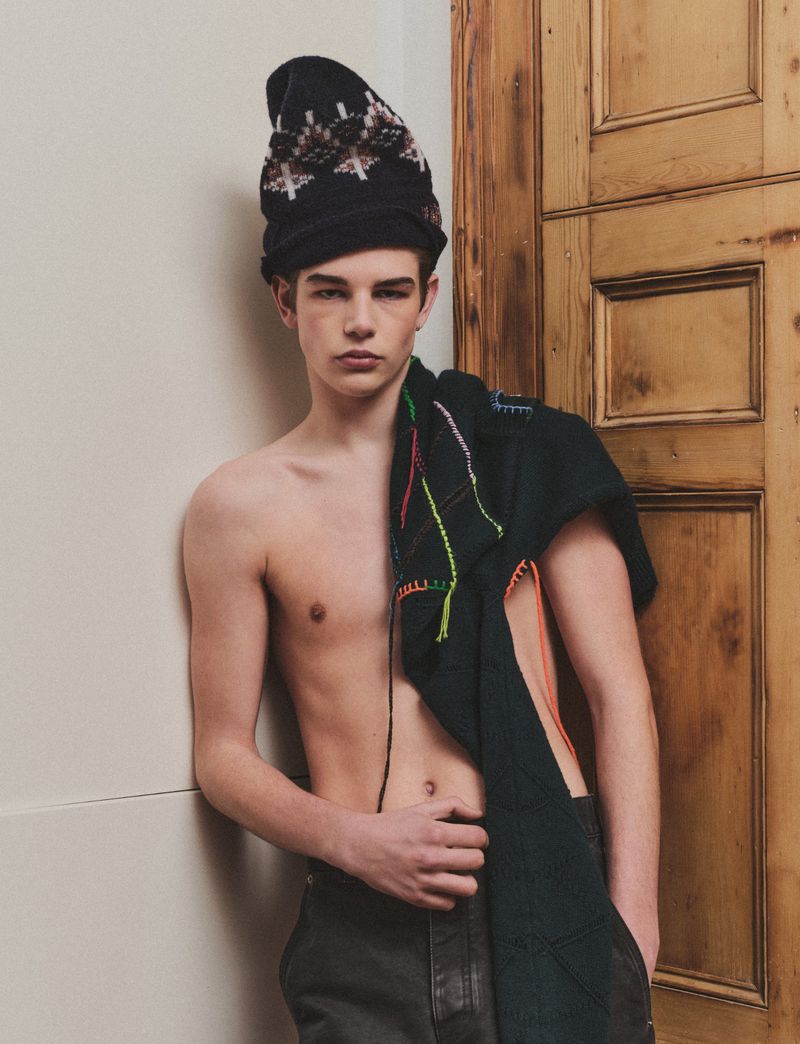 Young man wearing a black patterned beanie and dark trousers, with a dark knitted cloth draped over his shoulder, leaning against a wood door.
