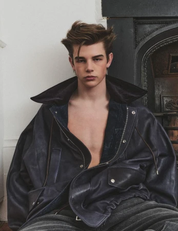 Portrait of a young adult male model with tousled brown hair wearing an unzipped navy blue leather jacket indoors with a dark fireplace.
