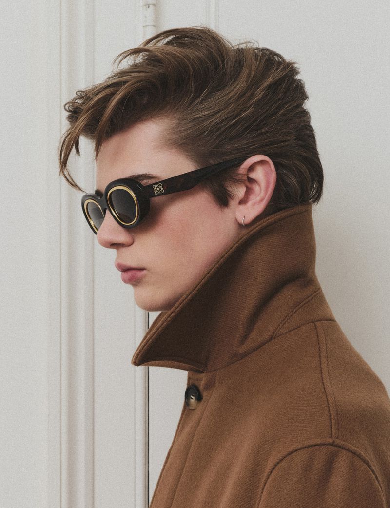 Portrait of a young adult male model with brown hair and sunglasses wearing a brown overcoat against a white panel background.