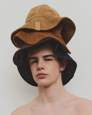 Portrait of a young man wearing three brown bucket hats stacked on his head in a studio setting with a neutral background.