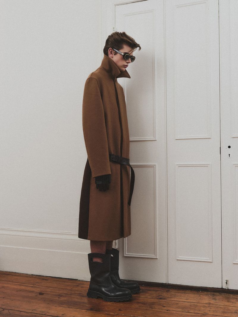 Profile of a young male model wearing sunglasses, a long brown belted coat, and black boots against white paneled doors.