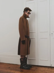 Profile of a young male model wearing sunglasses, a long brown belted coat, and black boots against white paneled doors.