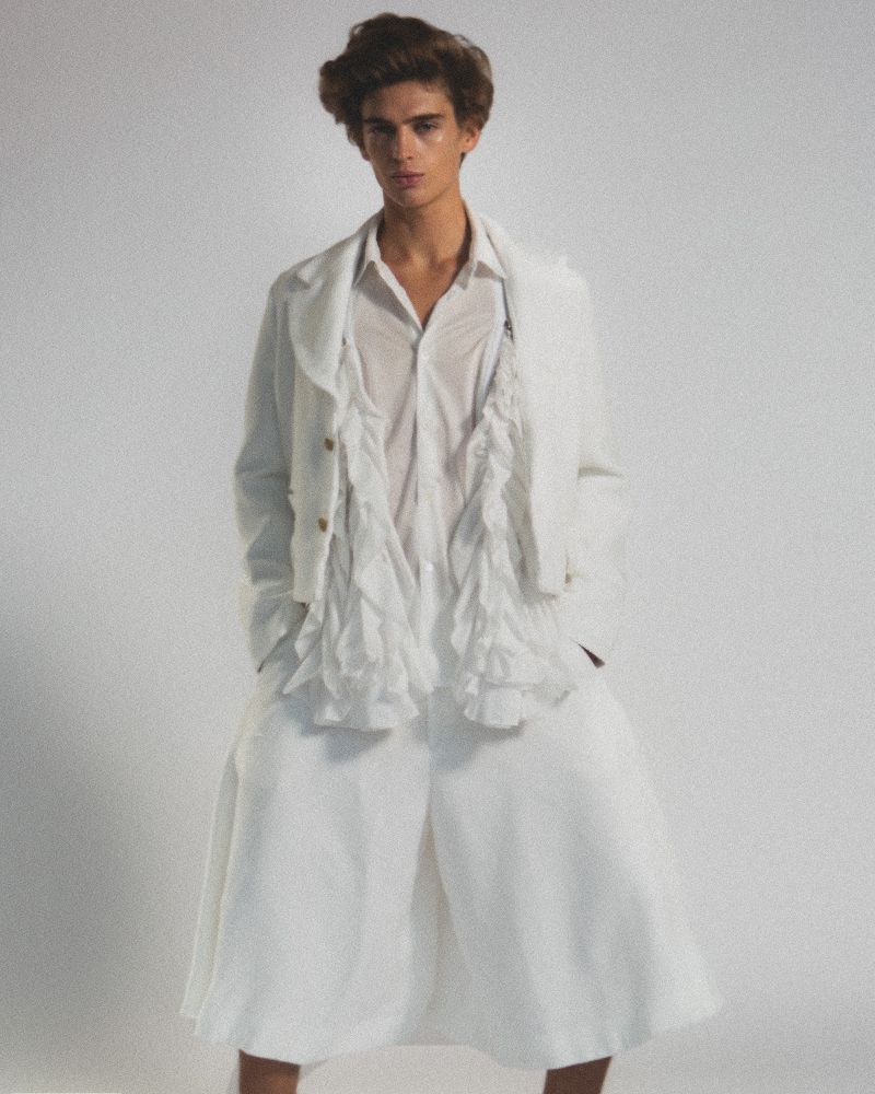 A young adult man poses in wide white culottes, a partially open white shirt with ruffles, and a matching jacket against a plain background.