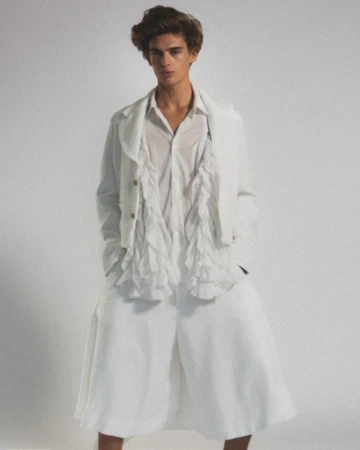 A young adult man poses in wide white culottes, a partially open white shirt with ruffles, and a matching jacket against a plain background.