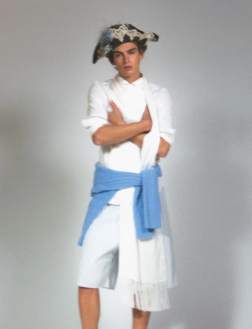 Male fashion model posing indoors wearing a white shirt, striped shorts, a light blue sweater, and a decorative pirate-style hat.