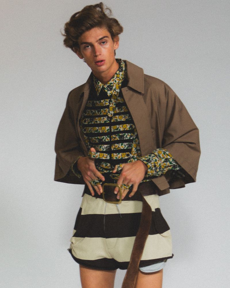 Fashionable male model in studio, wearing floral shirt, short jacket, striped shorts, belt. Neutral backdrop, portrait orientation.