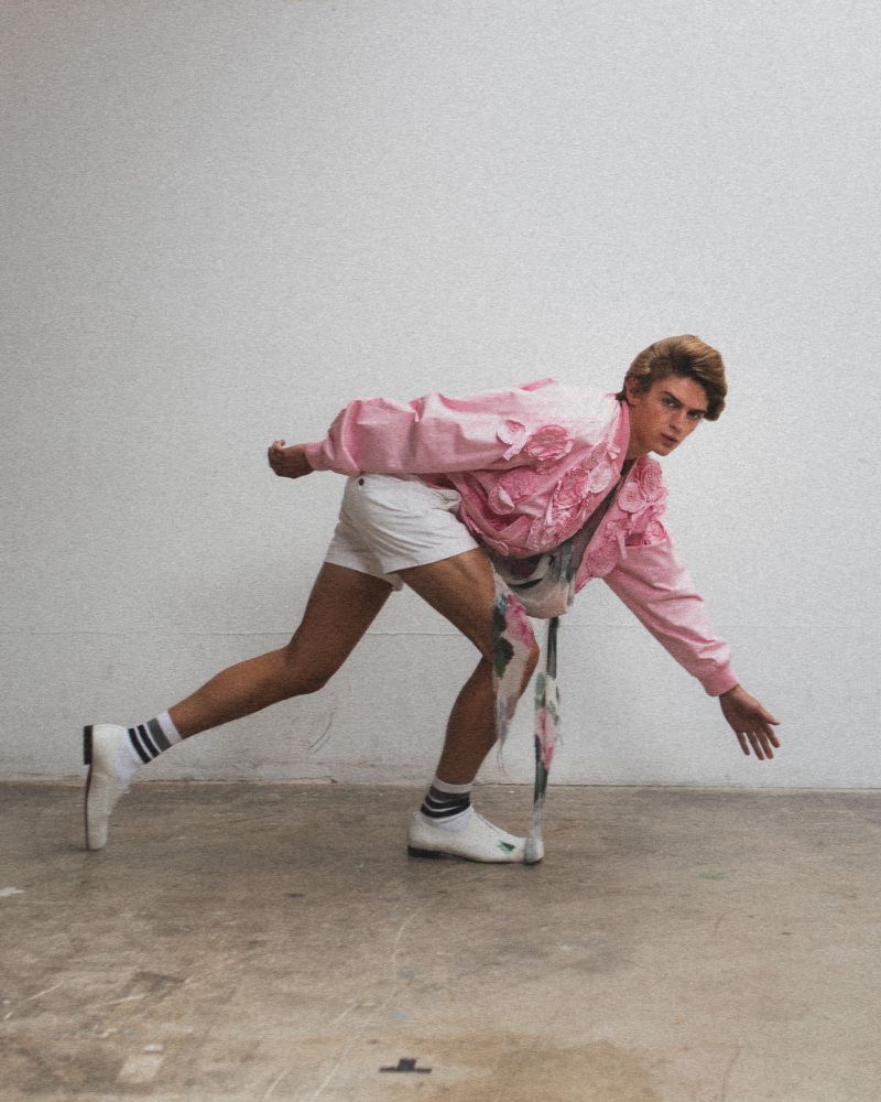 Young adult in pink jacket and white shorts strikes a dynamic pose against a white backdrop, fashion-forward and energetic.