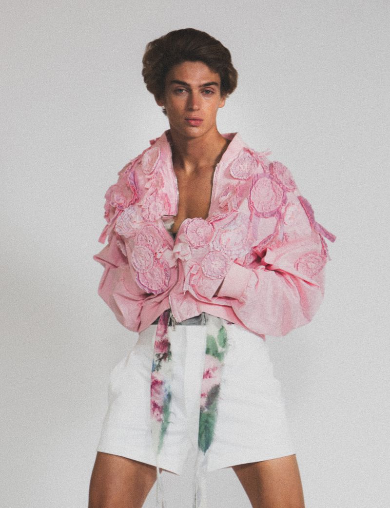 Young adult male poses in a light pink jacket with flower accents and white shorts with a floral belt against a white background.